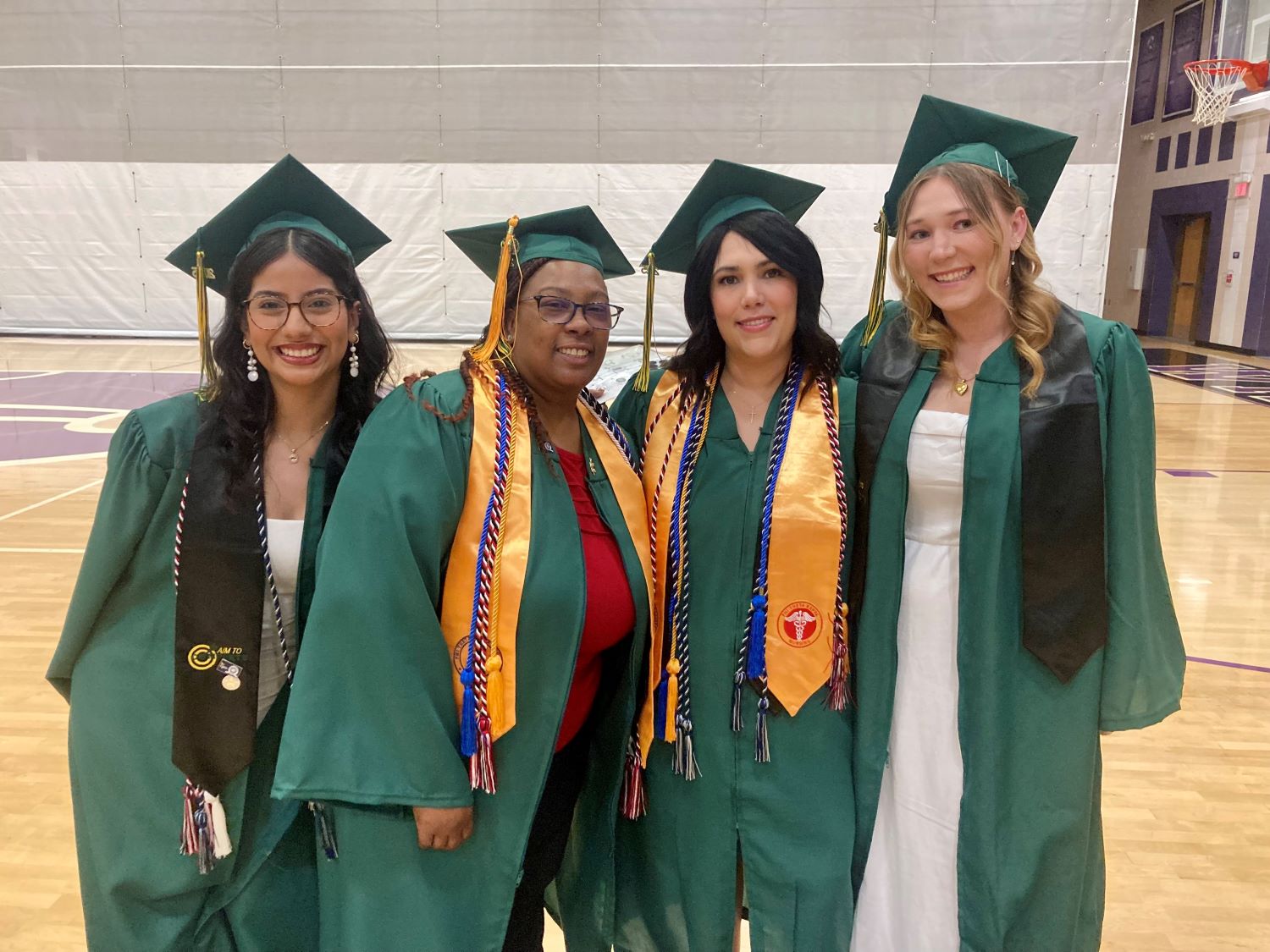 four women wearing caps and gowns, posing in front of the camera in the gymnasium