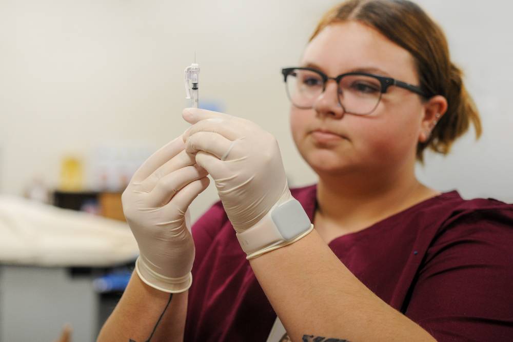 female allied health student wearing scrubs, rubber gloves, and holding a syringe 