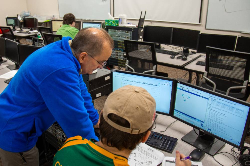 teacher standing next to seated student in front of computer monitors