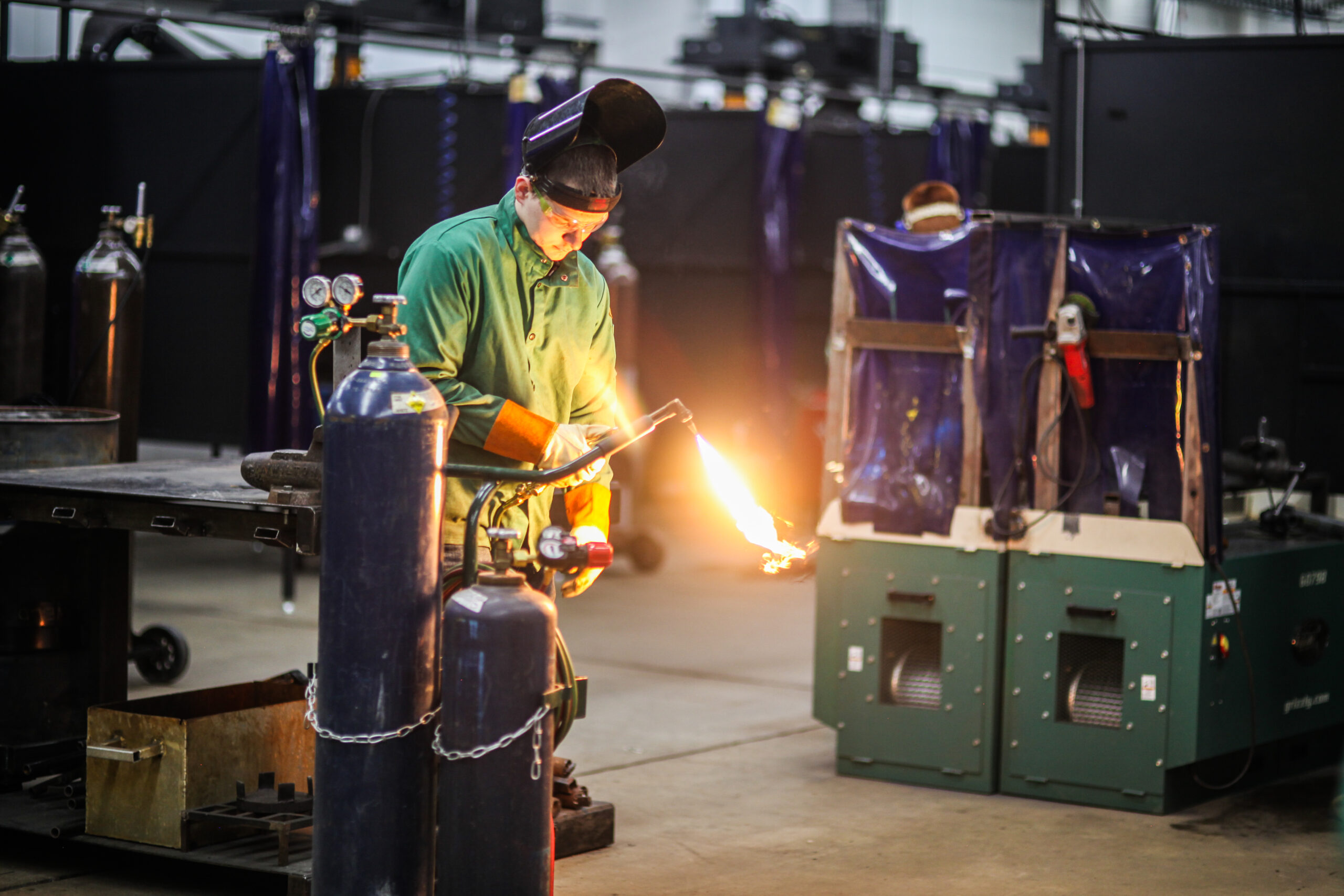 male student wearing green welding smock gloves, holding a torch, and wearing a welding mask that is lifted up to see his face