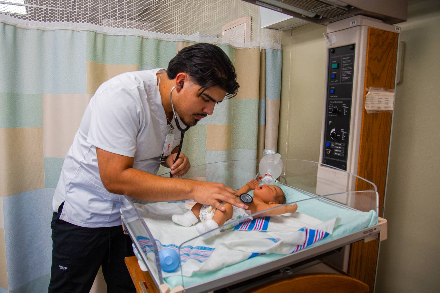 male nursing student wearing white nursing scrub and black pants, using a stethoscope to listen to the heartbeat of an infant