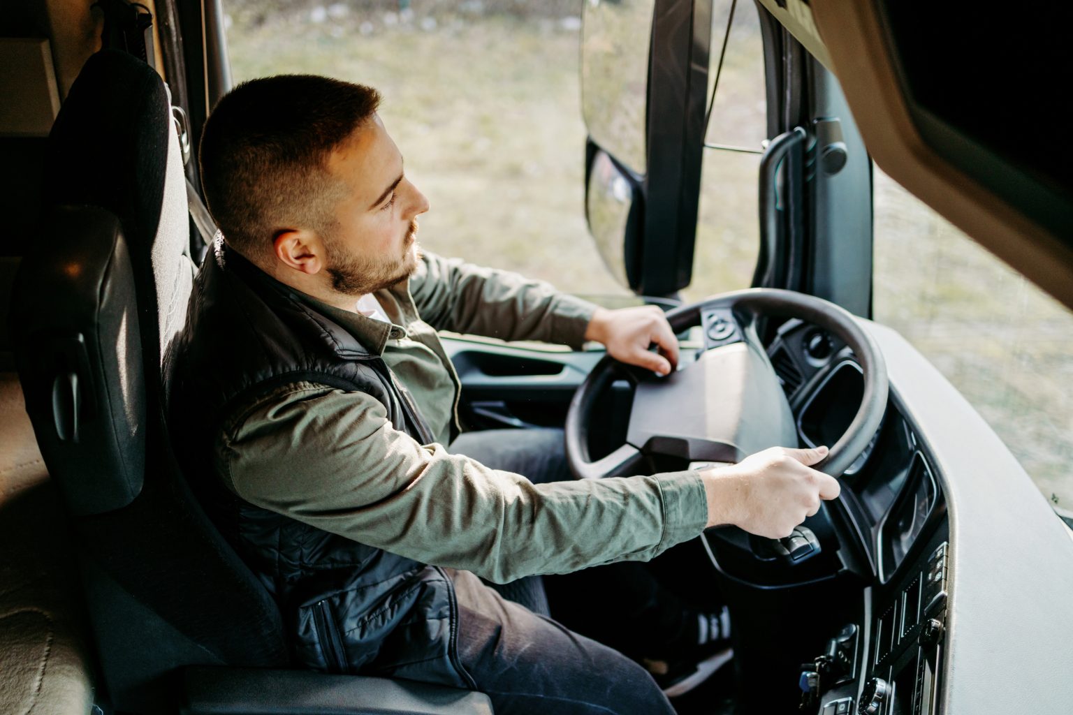 man sitting in the cab of a semi truck hands on the steering wheel and looking out the window.