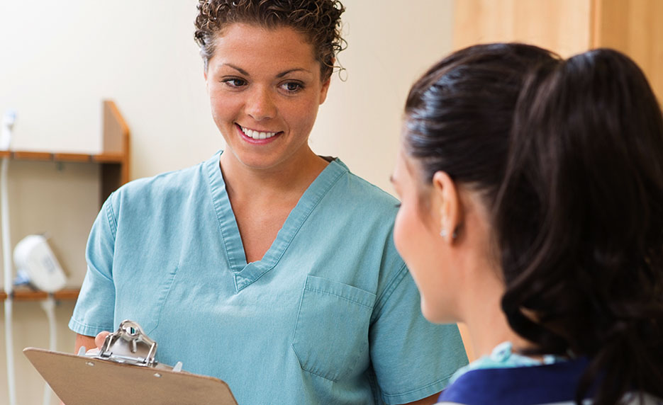 Female in scrubs, holding clipboard and smiling as she's talking to a patient