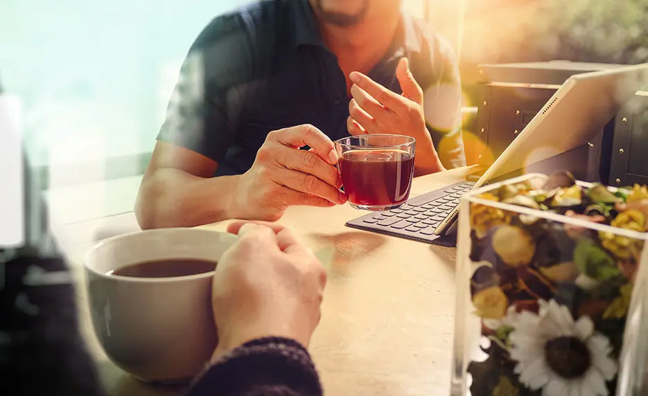 Close up of person from the shoulders down holding a cup of tea, sitting across from another person. All you see is their hand also holding a cup of tea.