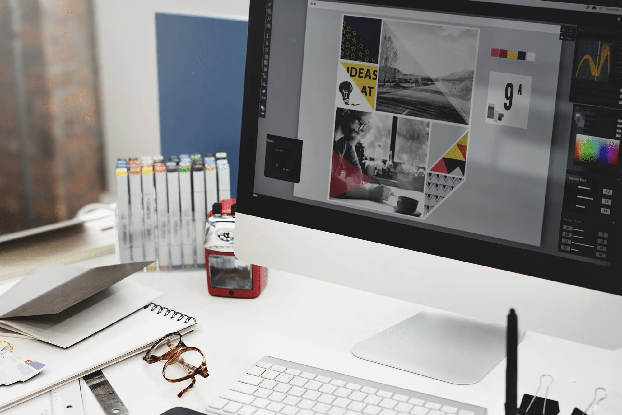 Keyboard and computer monitor sitting on a table with a notebook, a pair of eyeglasses, and some colorful writing utensils