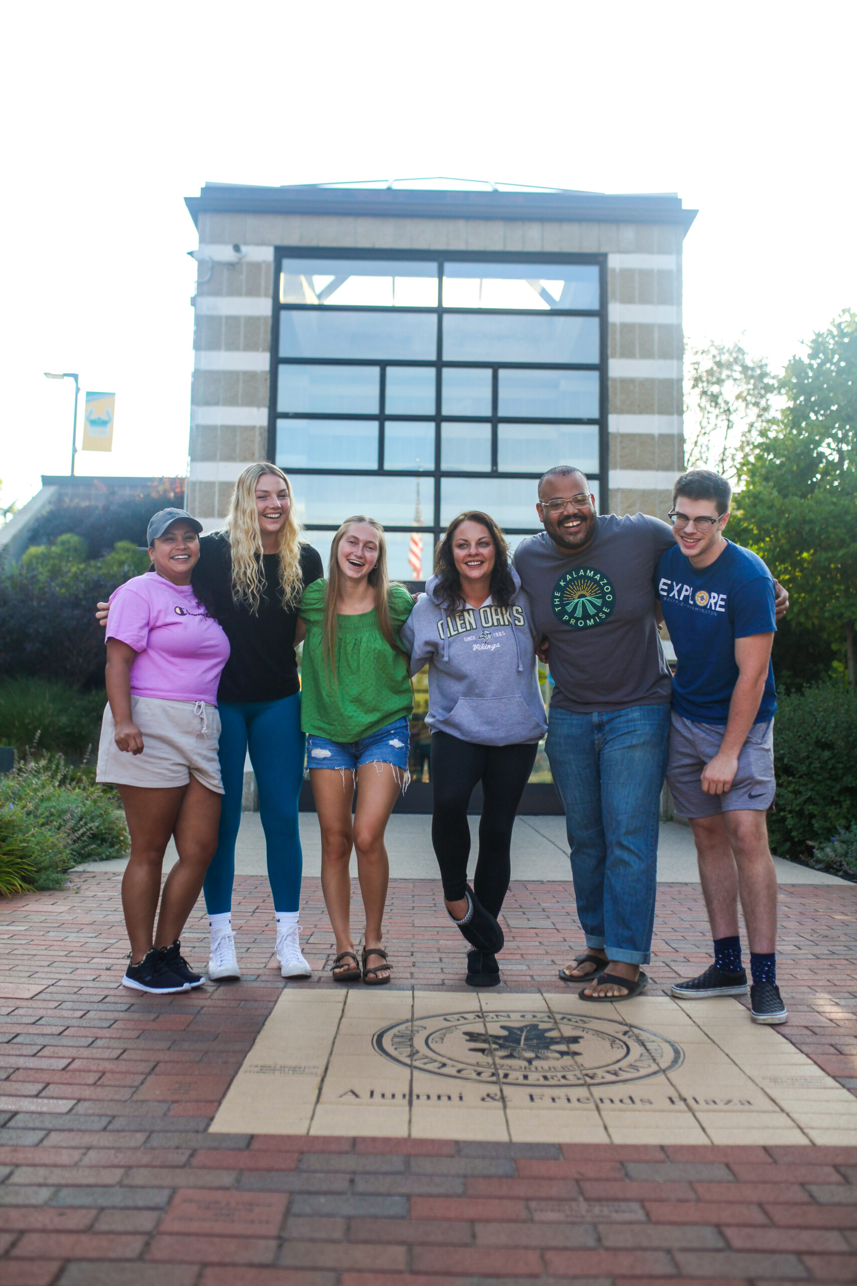Six students of different ages and genders standing arm-in-arm in front of the main entrance of Glen Oaks Community College.