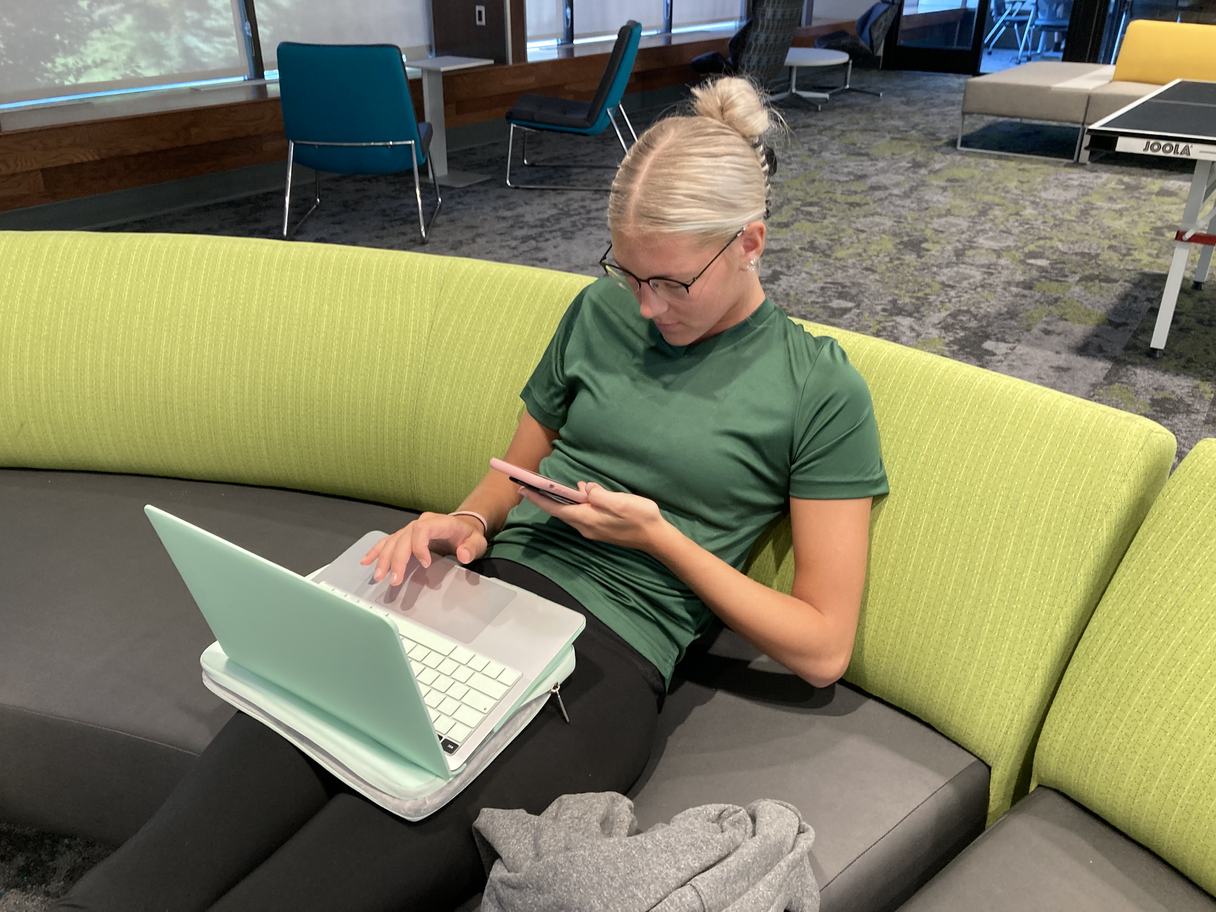 female student sitting on couch with laptop on her lap and looking at her phone