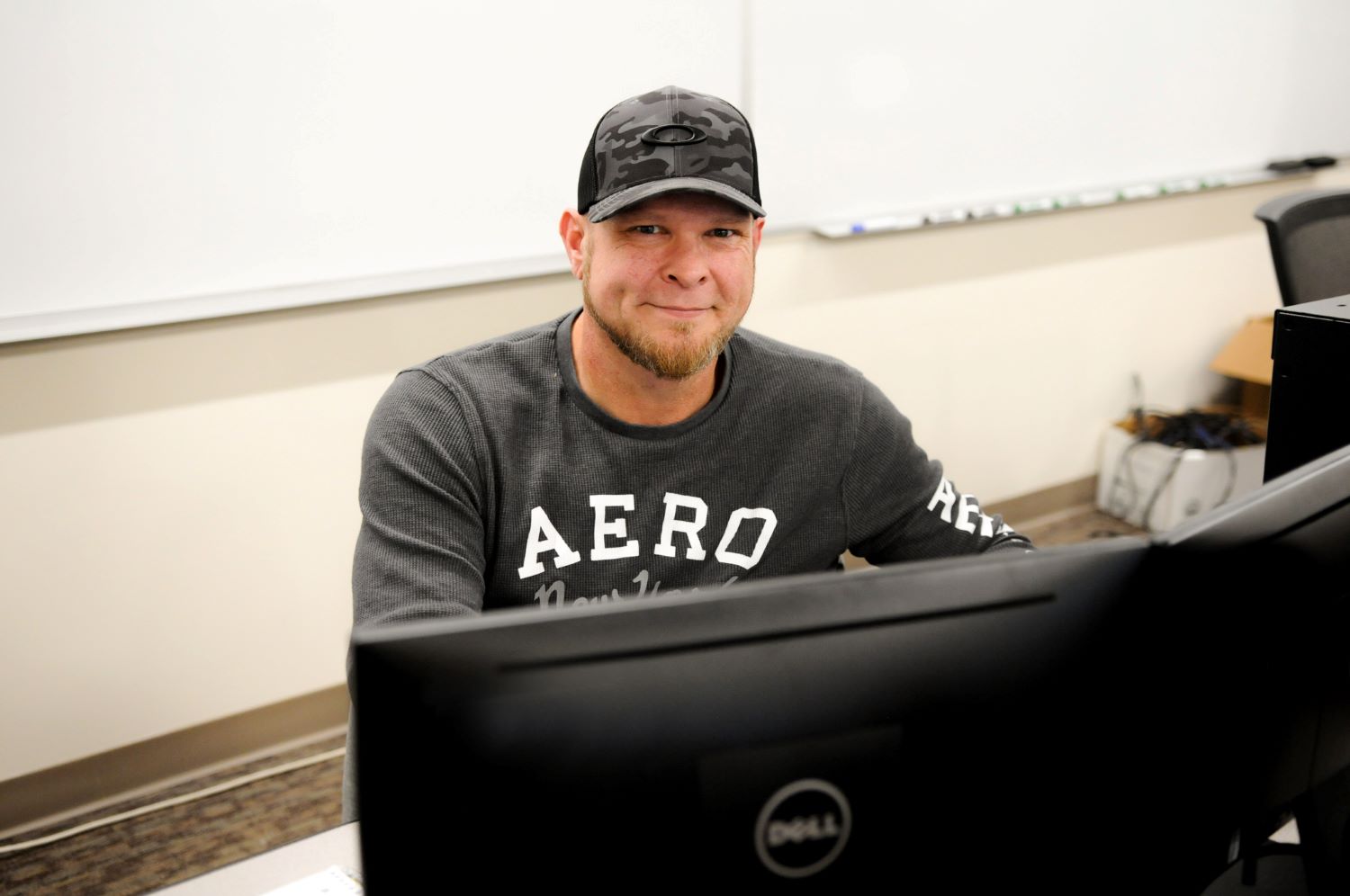 Male student wearing a gray shirt and gray baseball cap, smiling for the camera, sitting in front of computer screens